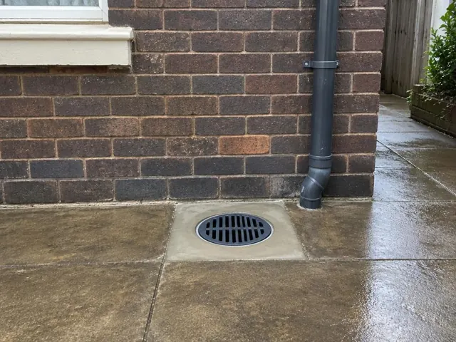 Clean outdoor drain grate set in wet paved area beside a brick wall in Tighes Hill
